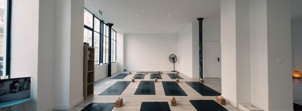 Spacious Lomey Yoga Studio in Paris with black mats neatly aligned on a light wooden floor. Sunlight streams through large windows, creating a serene atmosphere.