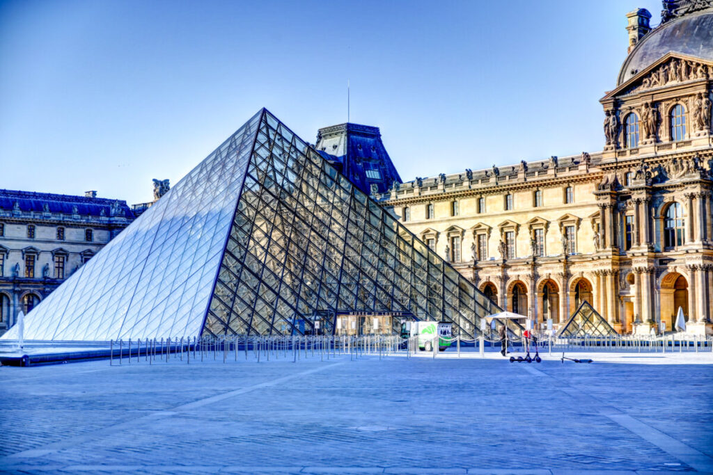 The Louvre Pyramid, a large glass structure, in front of the historic Louvre Museum with ornate architecture against a clear blue sky.