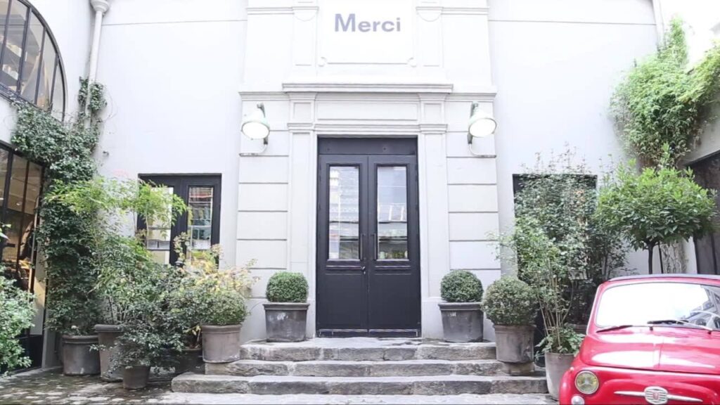 Entrance of Merci Shop a charming boutique labeled “Merci” with a black door, surrounded by lush greenery in pots in Paris