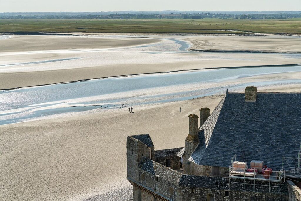 Medieval stone building with a slate roof overlooks a vast, sandy coastal landscape with a winding river and distant greenery under a clear sky.