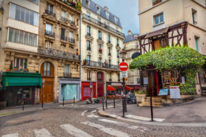 Montmartre street with historic buildings, ornate balconies, and vibrant awnings. A tree-covered café, scooters, and a "no entry" sign add depth.