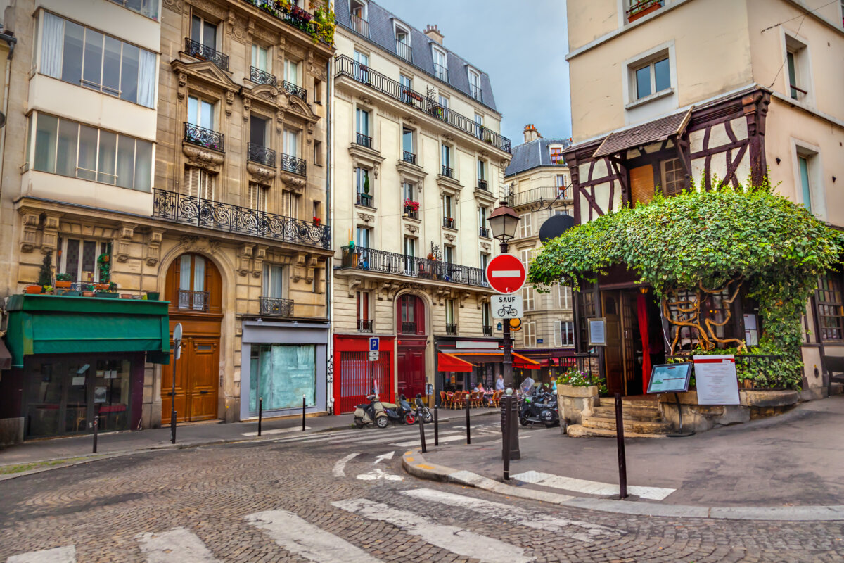 Montmartre street with historic buildings, ornate balconies, and vibrant awnings. A tree-covered café, scooters, and a "no entry" sign add depth.