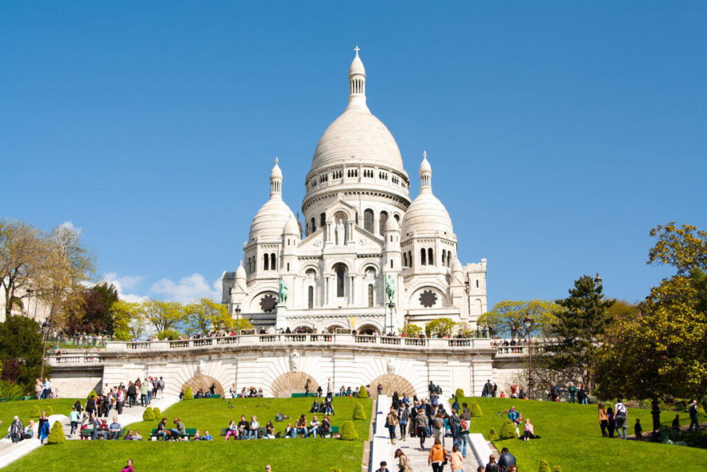 Montmartre Sacré-Cœur Basilica on a sunny day, with people relaxing on vibrant green grass. Majestic architecture against a clear blue sky.