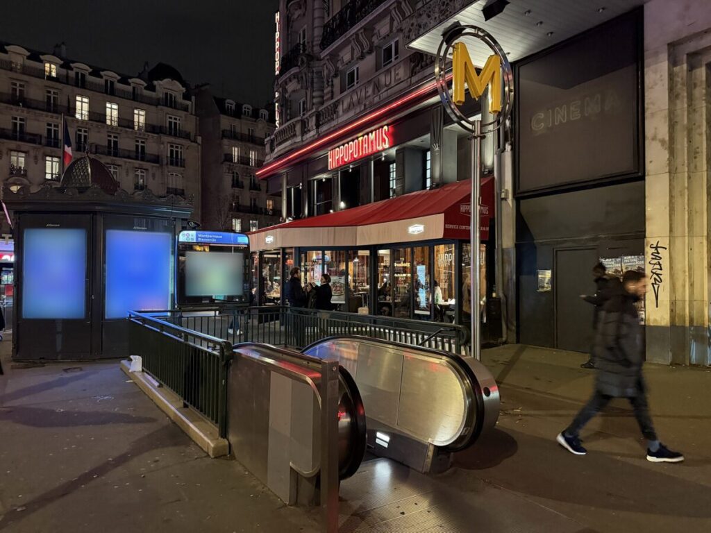 Nighttime view of Montparnasse Station in Paris, featuring pedestrians strolling along a lively city street.