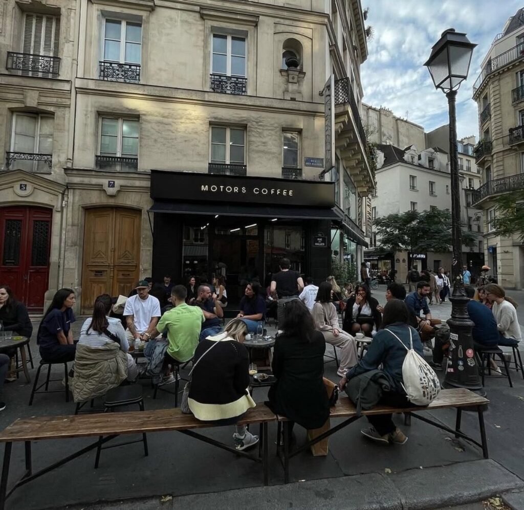 Bustling outdoor Motors Coffee cafe scene with people sitting on benches and chairs in front of a classic Parisian building. The atmosphere is lively and social.
