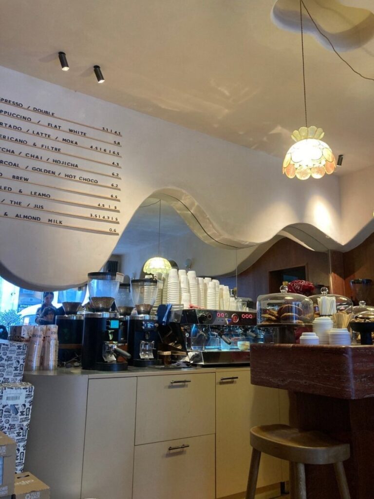 Noir - Coffee Shop & Torréfacteur interior with a wavy mirror and menu board on the wall. Counter features coffee machines, cups, and baked goods under soft, warm lighting.
