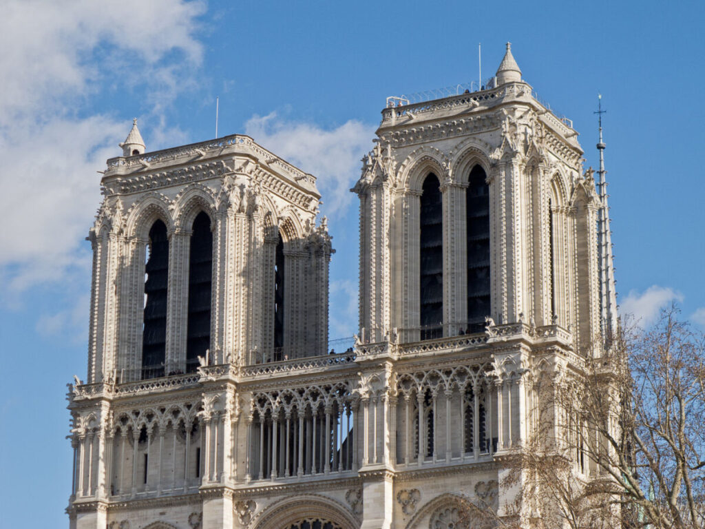 Twin Gothic towers of Notre-Dame Cathedral against a bright blue sky, detailed with arched windows and ornate stone carvings, evoke timeless elegance.