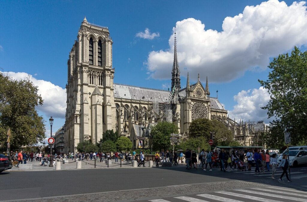 Notre Dame Paris with tall spires and detailed stonework under a blue sky with clouds. Busy street in front with people walking and trees nearby.