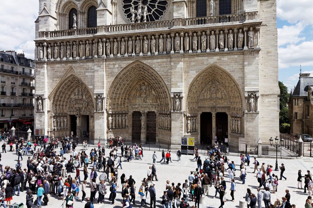 Notre Dame Cathedral facade with arched doorways and intricate stone carvings. Busy scene with tourists on a sunny day in Paris.
