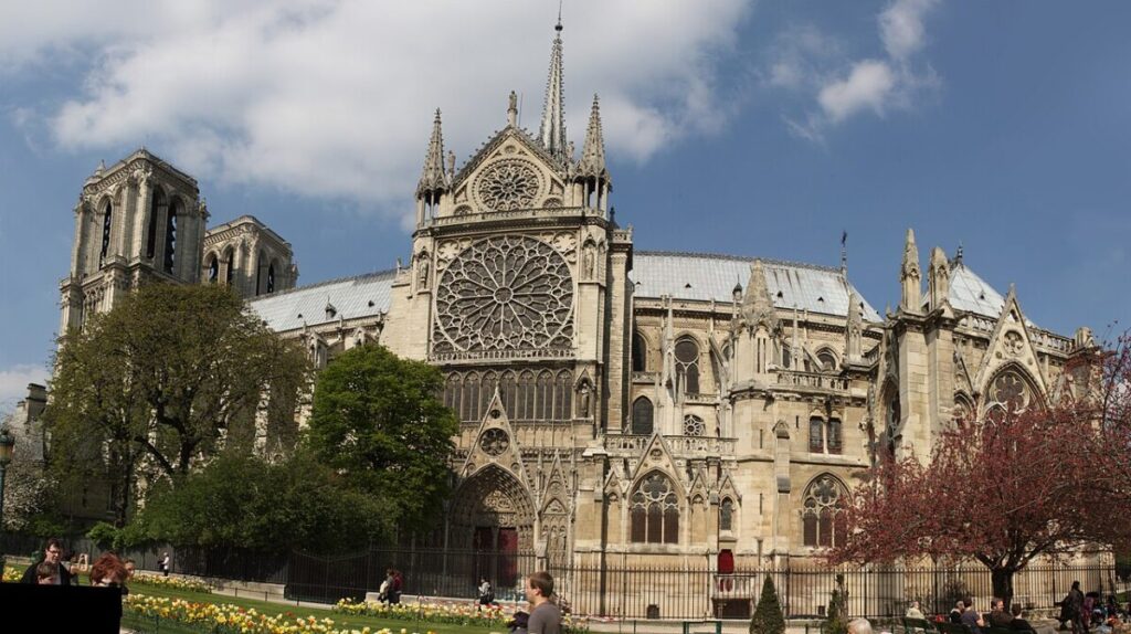 Notre Dame Paris with intricate rose window, ornate facade, and stone spires under a clear sky. Trees and people in a vibrant garden foreground.