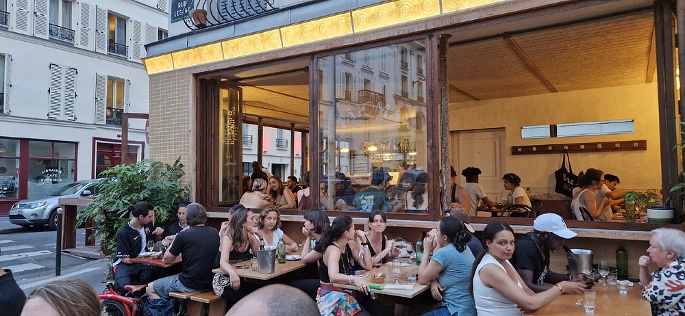 People sit at tables outside Paloma, Paris with large windows on a corner street.