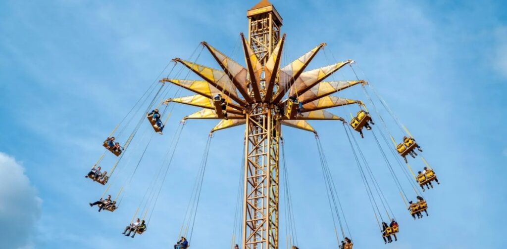 Parc Astérix Tour Numerobis tall, golden swing ride at an amusement park ascends against a blue sky. People in chairs are swinging outward, expressing excitement and adventure.