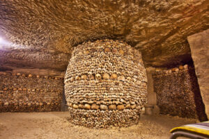 Haunting underground space in the Paris Catacombs, filled with dim light and walls of stacked human skulls and bones.