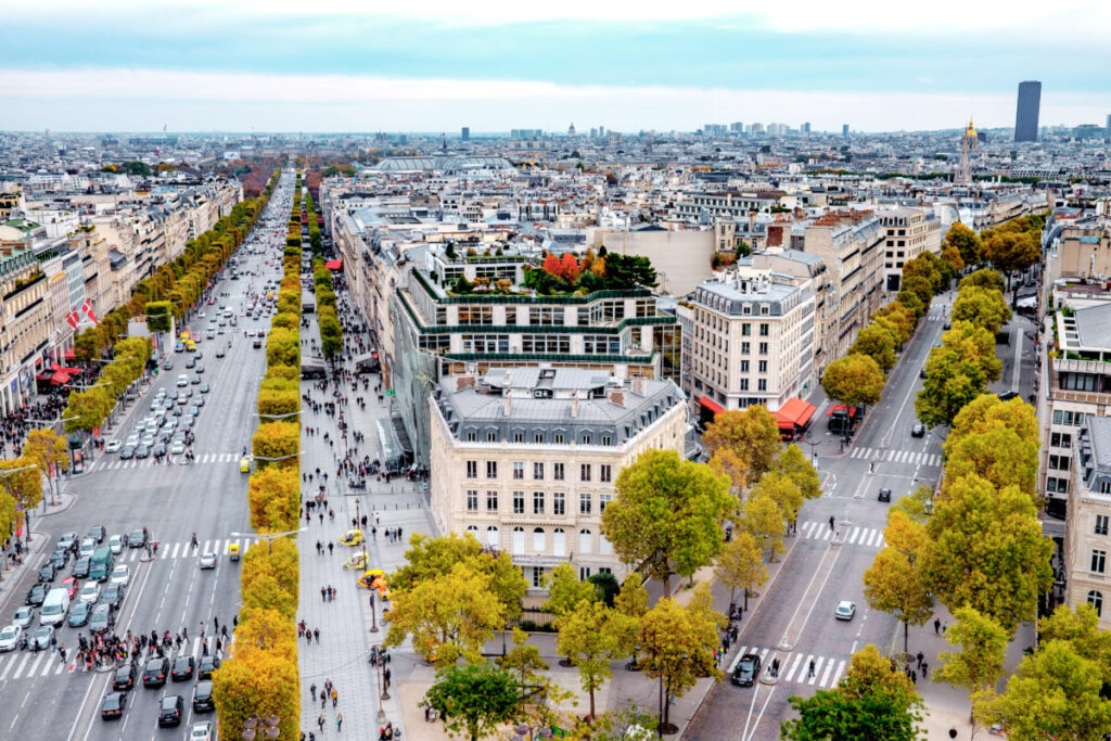 Aerial view of a bustling Parisian intersection, featuring tree-lined avenues and classic European buildings under a partly cloudy sky. The scene is vibrant and lively.