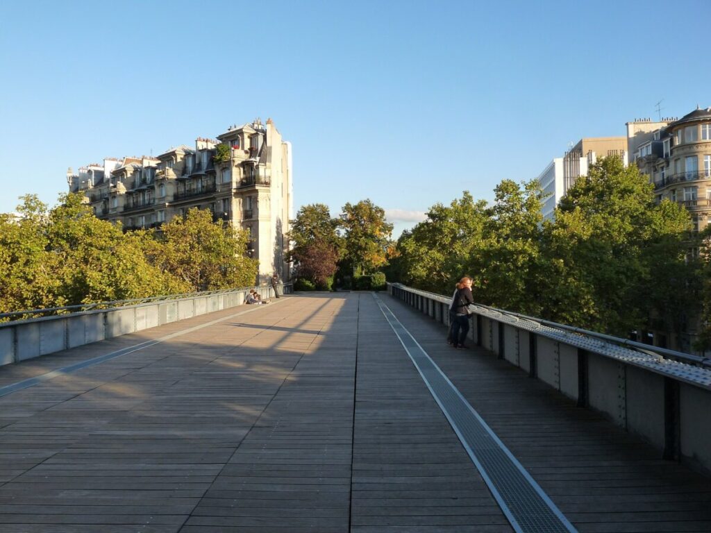 Individual strolling along the Coulée Verte René-Dumont bridge, with a view of the Paris city skyline.