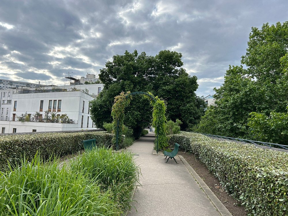 Scenic pathway with lush green bushes and a white building, set in the Paris Coulée Verte René-Dumont Garden.