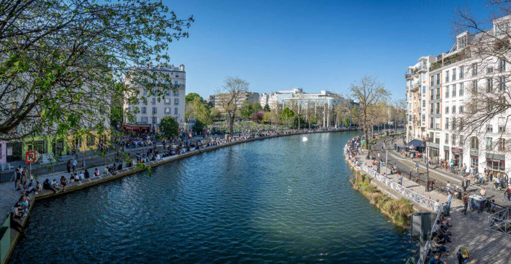 Panoramic view of people sitting near white curved buildings, colorful stores, a bridge, and trees in Paris, France.