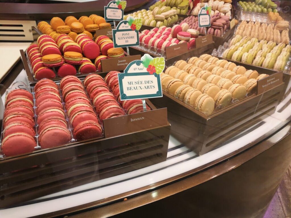 Display case filled with vibrant macarons in shades of red, orange, pink, and beige. Signs with names add a touch of elegance and sophistication.