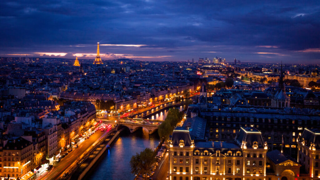 Aerial view of Paris at dusk, featuring the illuminated Eiffel Tower and city lights reflecting on the Seine River. The sky is dark with a purple hue.