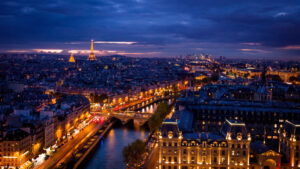 Aerial view of Paris at dusk, featuring the illuminated Eiffel Tower and city lights reflecting on the Seine River. The sky is dark with a purple hue.