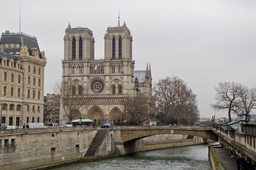 Notre-Dame Cathedral in Paris on a cloudy day, with the Seine River and an arched bridge in the foreground, surrounded by trees and historic buildings.