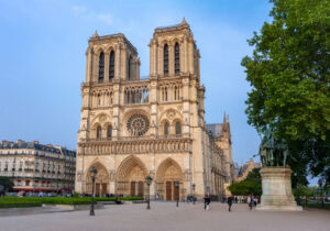 Notre-Dame Cathedral in Paris is shown at sunset, featuring twin towers, a large rose window, and Gothic architecture. People and a statue are in the foreground.
