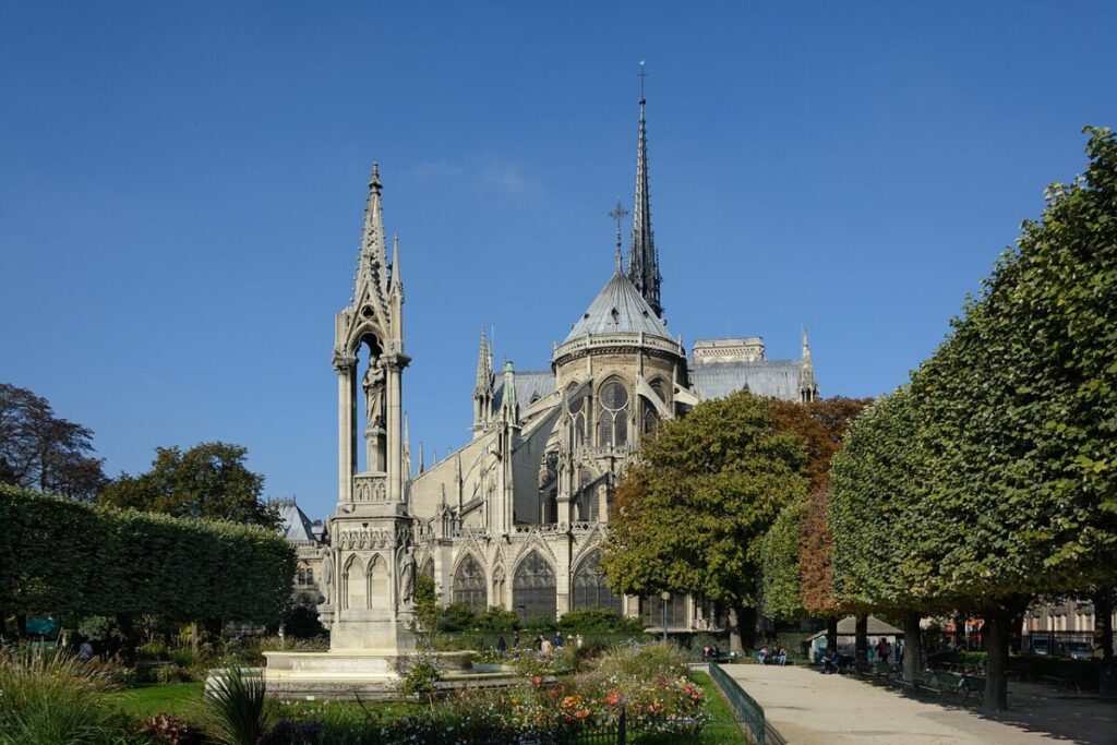 Paris Notre-Dame East with a tall spire, surrounded by lush trees and a manicured garden under a bright blue sky, conveying a serene and majestic atmosphere.