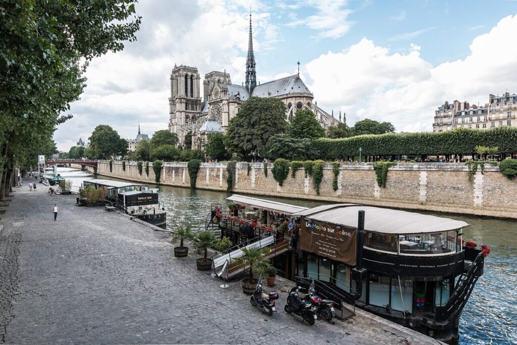 River scene with a historic cathedral and a tree-lined promenade. Houseboats with outdoor dining float on the river, under a partly cloudy sky.