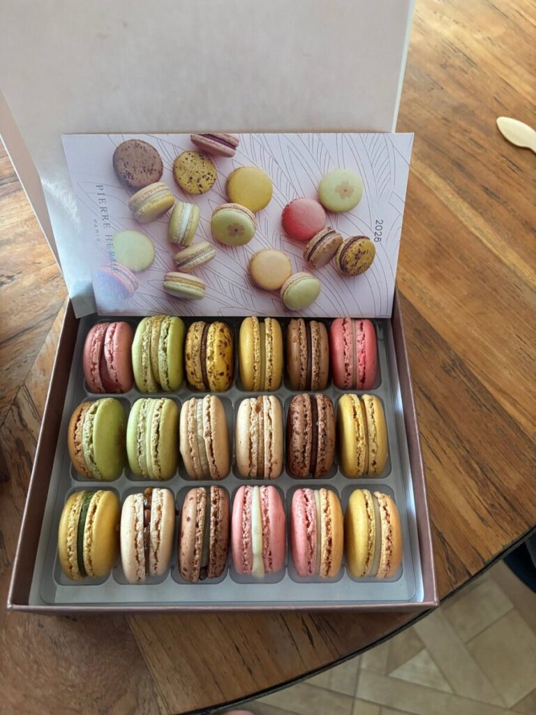 Box of Paris Pierre Hermé assorted macarons arranged neatly in rows on a wooden table. The macarons are pastel-colored with a small leaflet showing different flavors.