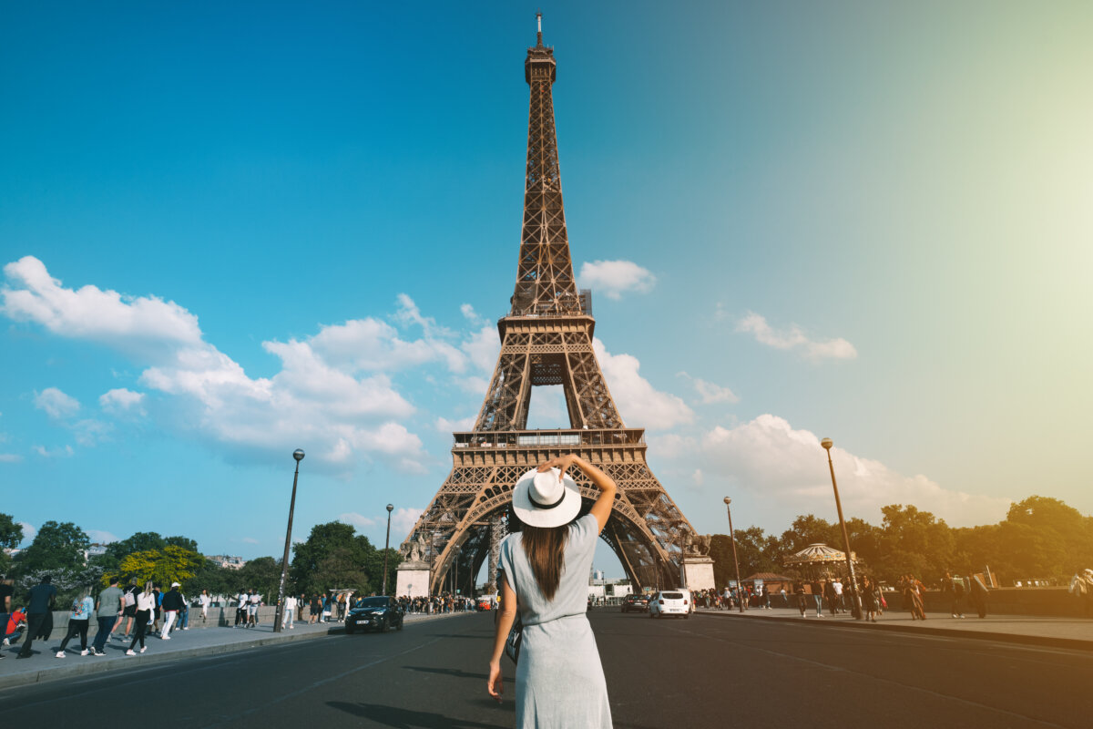 Woman in a sun hat stands with her back to the camera, facing the Eiffel Tower in Paris, embodying a travel vibe.