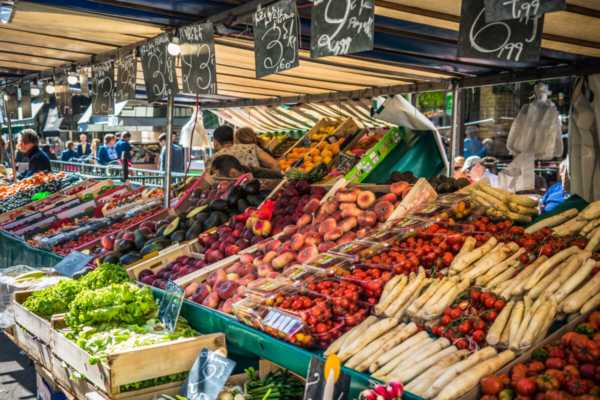 Pairsian Market bustling outdoor market stall showcasing fresh tomatoes, peaches, and asparagus under a shaded canopy with price signs.