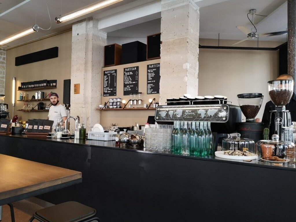 Partisan Café Artisanal shop interior with a barista behind a sleek counter. Espresso machine, glassware, snacks, and a menu board create a welcoming atmosphere.
