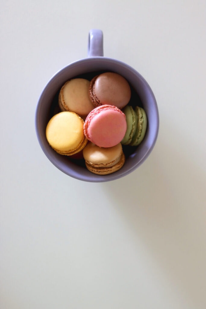 Purple mug filled with colorful macarons: pink, yellow, brown, and green, arranged on a light surface, exuding a playful and appetizing vibe.