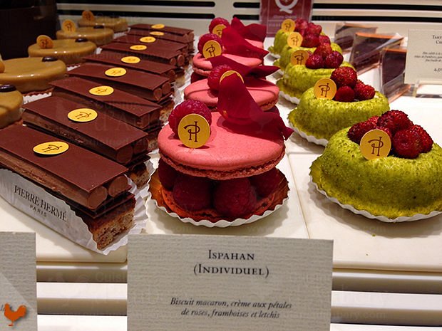 Display of elegant pastries featuring chocolate slices, pink macarons with raspberries, and pistachio cakes topped with raspberries in Pierre Hermé, Paris