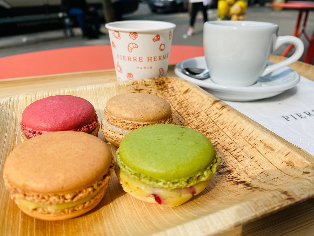 Pierre Hermé close-up of a wooden tray with colorful macarons in green, pink, and beige. Behind, there's a branded coffee cup and a saucer on a café table. The setting feels inviting and Parisian.