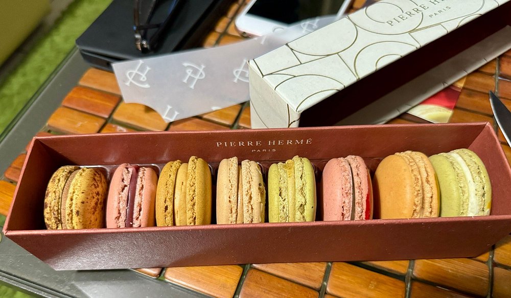 Box of assorted Pierre Hermé macarons in vibrant colors like brown, pink, green, and peach rests on a wooden table. A white box and smartphone are in the background.
