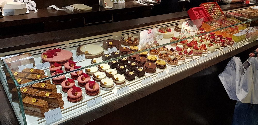 Pierre Hermé, Paris display case filled with various colorful pastries and cakes, including macarons, tarts, and layered desserts. The vibrant, tempting treats evoke a sense of indulgence and sophistication.