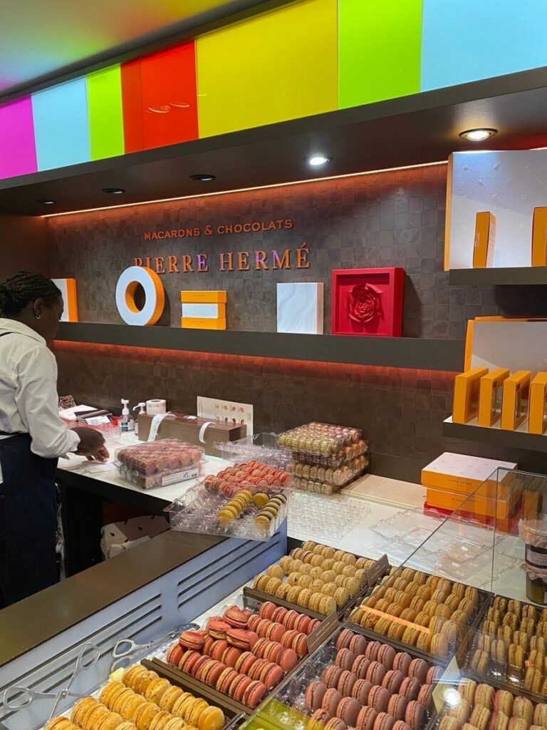 Pierre Hermé colorful patisserie with a display of varied macarons in glass cases. A staff member packages treats, set against a vibrant, modern backdrop.