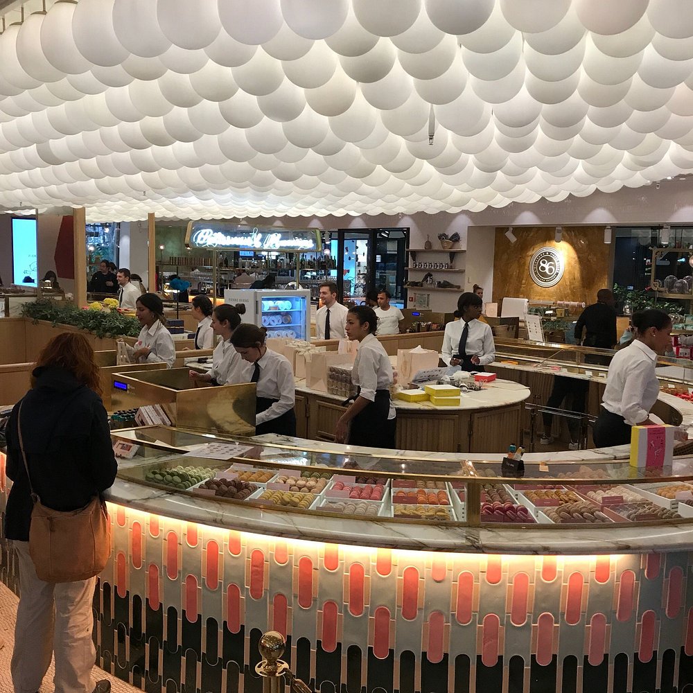 Pierre Hermé, Paris, with vibrant lighting and a cloud-like ceiling. Staff in white shirts assist customers at a colorful macaron display counter.