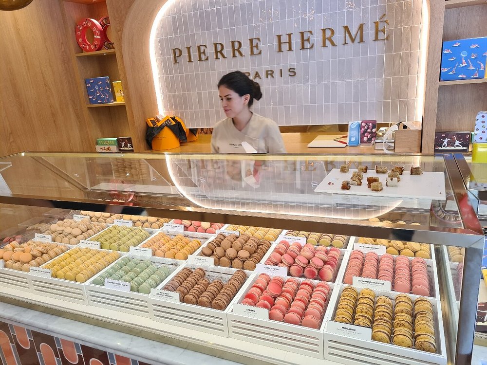 Employee stands behind a glass counter of Pierre Hermé displaying colorful macarons at Pierre Hermé, Paris. Shelves with boxes add a vibrant, upscale ambiance.