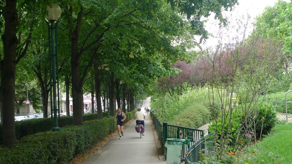 In the Promenade Plantée, Paris, a woman cycles down the sidewalk next to a man enjoying the scenery.
