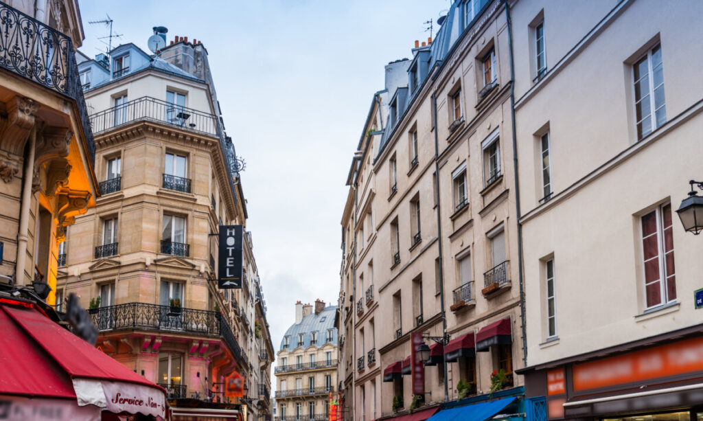 Charming Paris street scene with classic, ornate buildings on both sides. A hotel sign hangs on the left. The atmosphere is lively and inviting.