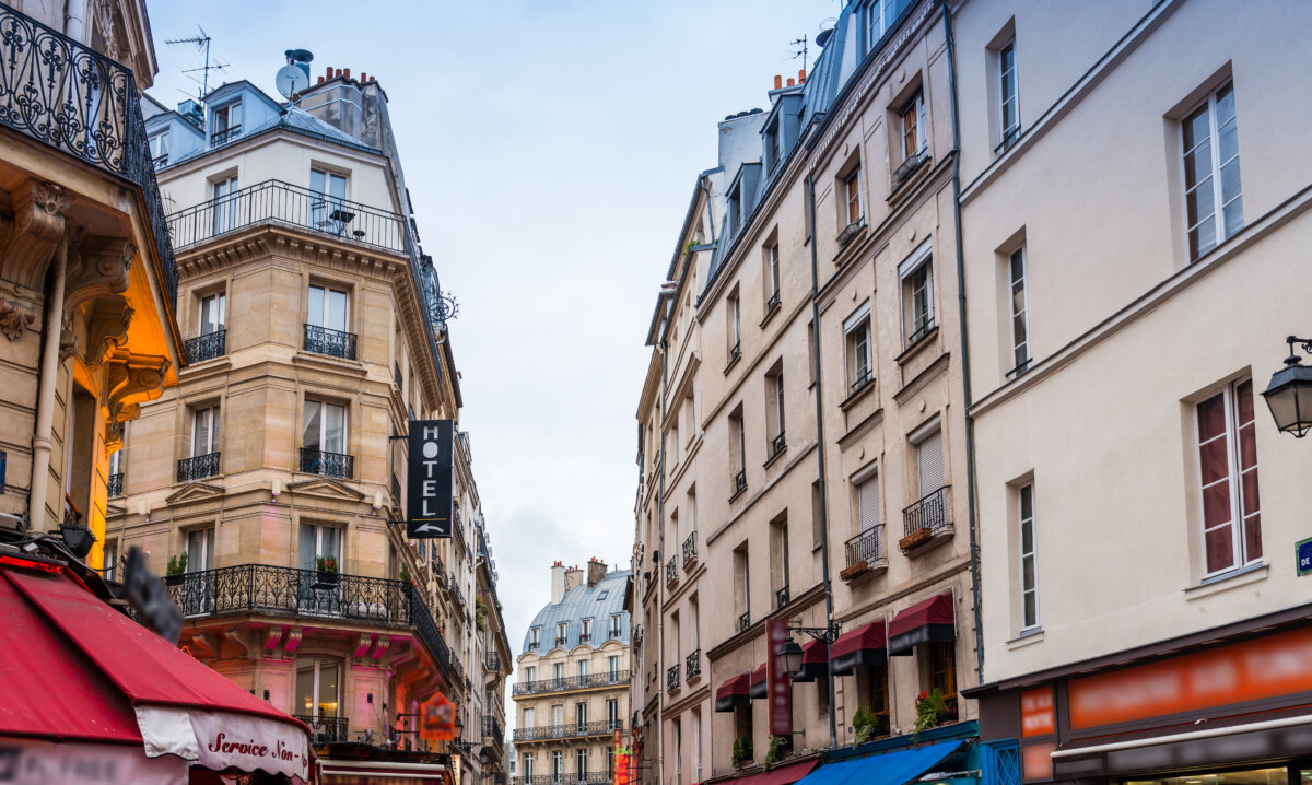 Charming Paris street scene with classic, ornate buildings on both sides. A hotel sign hangs on the left. The atmosphere is lively and inviting.
