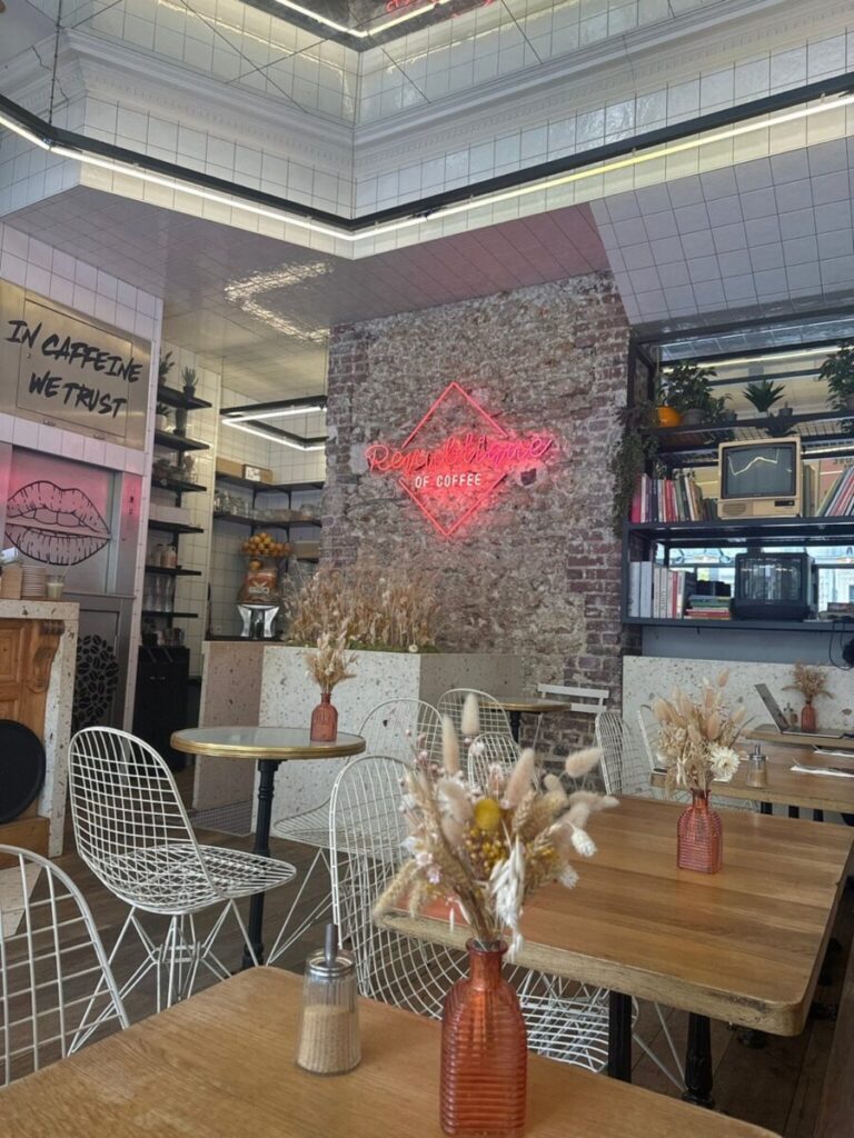République of Coffee café with wood tables, white wire chairs, and dried flowers in orange vases. Exposed brick wall with pink neon sign reads “République of Coffee.”
