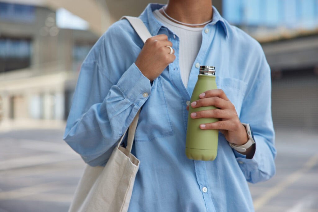 Person wearing a blue shirt holds a green bottle and white tote bag. They have a smartwatch on their wrist. Background features modern urban architecture.
