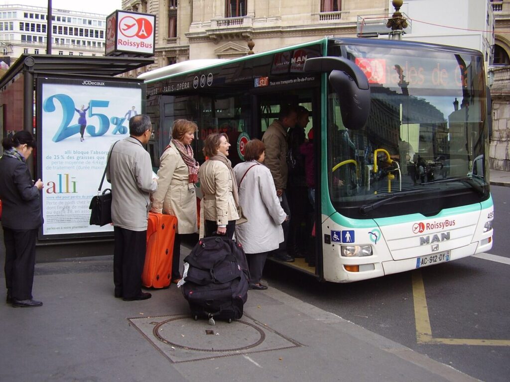 People in coats boarding a RoissyBus in an urban setting. Large suitcases are nearby, suggesting travel. The mood is busy and purposeful.