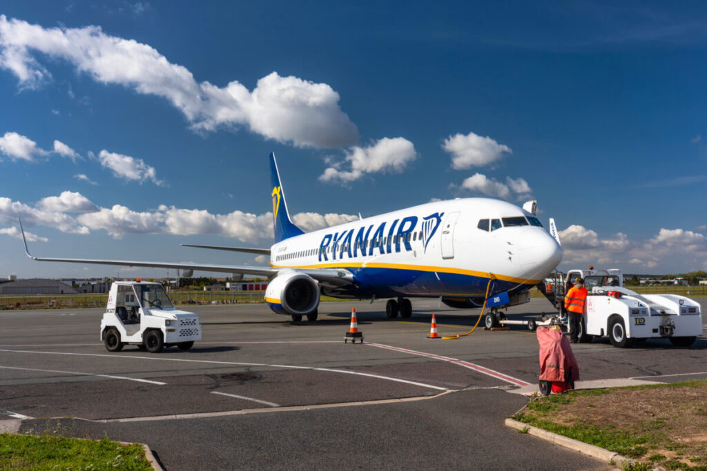 Ryanair plane parked at an airport in Paris, France, with terminal buildings and other aircraft in the background.
