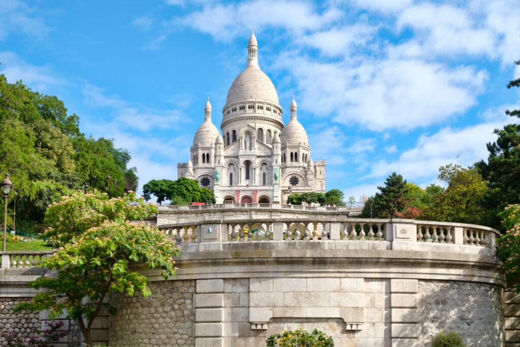 White domed Sacré-Cœur Basilica stands against a blue sky, surrounded by lush greenery and stone balustrade. The scene is serene and majestic.