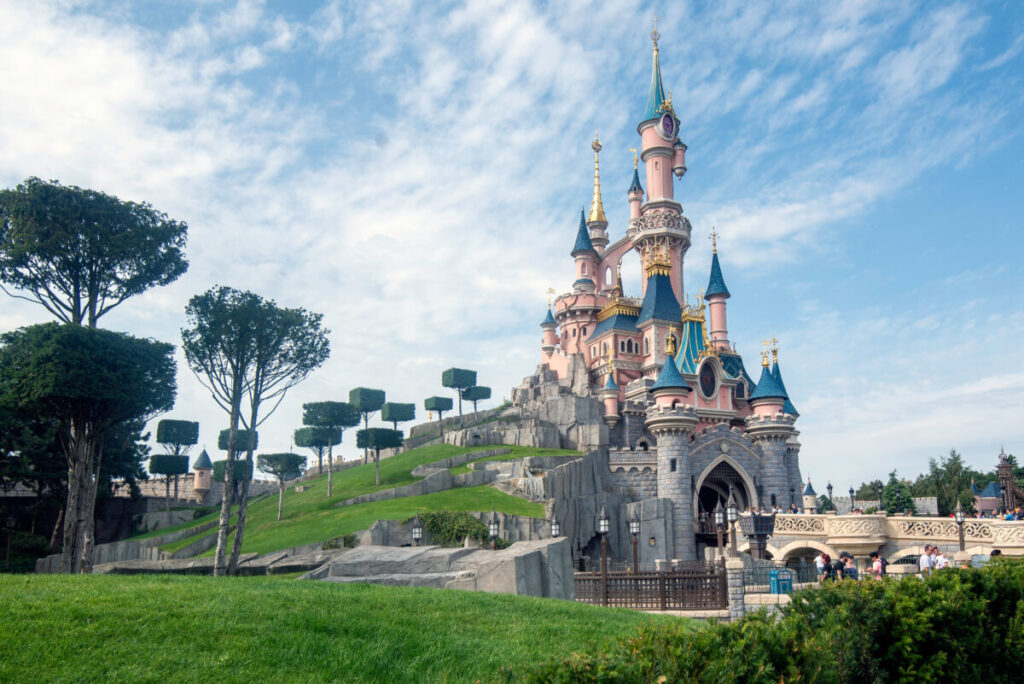 Sleeping Beauty Castle with pink towers and blue roofs, set on a lush green hill under a blue sky with wispy clouds in Disneyland, Paris