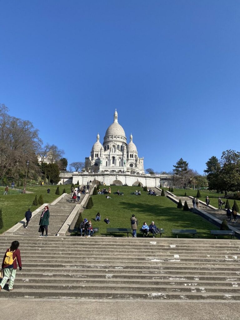 People relax on green lawns leading up to the Sacré-Cœur Basilica under a clear blue sky. The scene conveys a peaceful, leisurely atmosphere.
